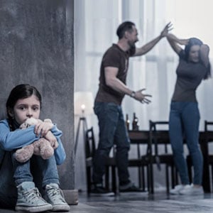 A child with a teddy bear observes her parents in a heated discussion near a window.